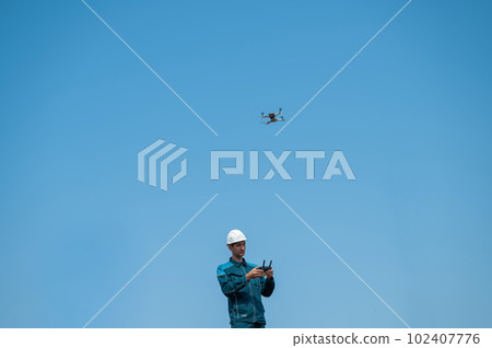 A man in a helmet and overalls controls a drone at a construction site. The builder carries out technical oversight. 102407776