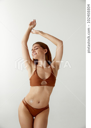 Portrait with beautiful young, smiling girl, woman in swimsuit using sunscreen cosmetic on body over white color studio background. Suncream, sunblock 102408804