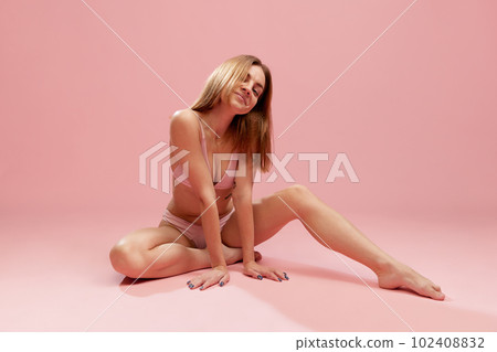 Portrait of young woman, girl wearing underwear sitting on floor and smiling over pink studio background. Model posing Portrait of young woman, girl wearing underwear sitting on floor and smiling over pink studio background. Model posing 102408832
