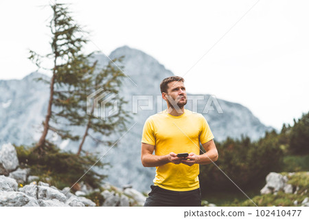 Waist up caucasian man in a yellow shirt looking up in the sky while holding phone in his hands, checking the weather forecast up in the mountains Waist up caucasian man in a yellow shirt looking up in the sky while holding phone in his hands, checking the weather forecast up in the mountains 102410477