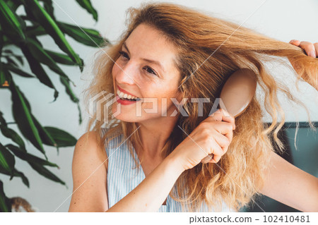 Waist up portrait smiling woman brushing her blonde curly hair with a hair brush  102410481