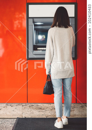 Vertical photo, back view of a woman standing in front of an ATM on a red wall, making a cash withdrawal Vertical photo, back view of a woman standing in front of an ATM on a red wall, making a cash withdrawal 102410483