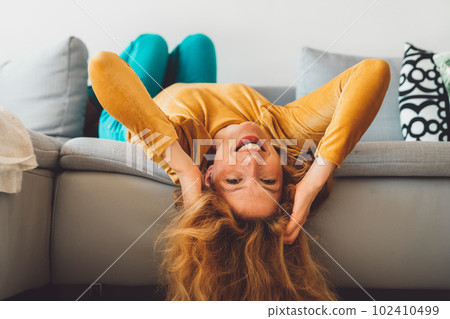 Smiling cheerful woman laying on the couch with her head hanging off, smiling at the camera wearing a red lipstick Smiling cheerful woman laying on the couch with her head hanging off, smiling at the camera wearing a red lipstick 102410499