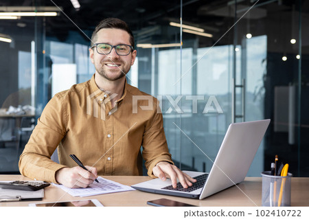 Portrait of young businessman in shirt, man smiling and looking at camera at workplace inside office, accountant with calculator behind paper work signing contracts and financial reports. Portrait of young businessman in shirt, man smiling and looking at camera at workplace inside office, accountant with calculator behind paper work signing contracts and financial reports. 102410722