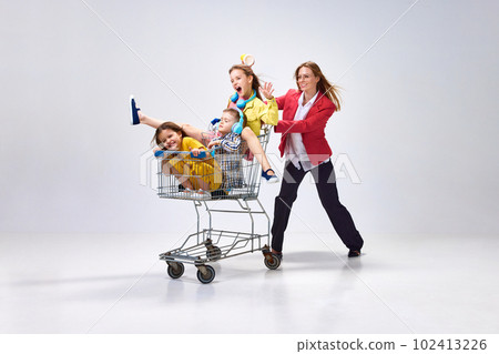 Portrait of happy young woman, mother going shopping with her little kids, children sitting in shopping trolley against grey studio background 102413226