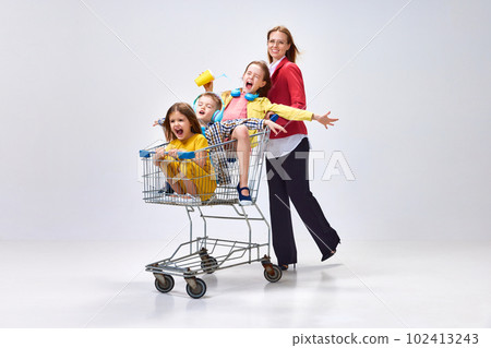 Portrait of happy young woman, mother going shopping with her little kids, children sitting in shopping trolley against grey studio background Portrait of happy young woman, mother going shopping with her little kids, children sitting in shopping trolley against grey studio background 102413243