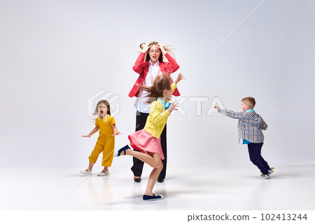 Portrait of tired young woman, mother holding head with hands around little noisy kids playing against grey studio background Portrait of tired young woman, mother holding head with hands around little noisy kids playing against grey studio background 102413244