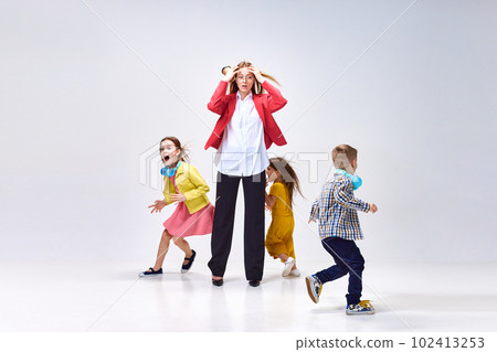 Portrait of tired young woman, mother holding head with hands around little noisy kids playing against grey studio background 102413253