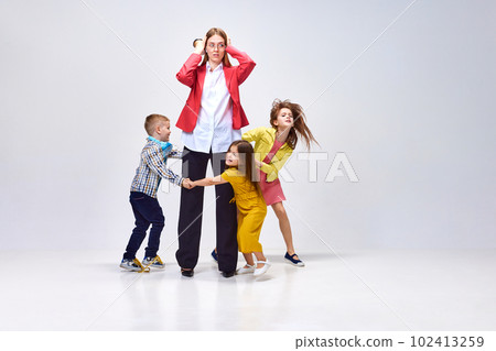 Portrait of tired young woman, mother holding head with hands around little noisy kids playing against grey studio background Portrait of tired young woman, mother holding head with hands around little noisy kids playing against grey studio background 102413259