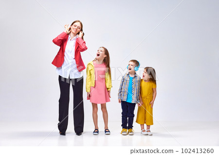 Rest needed. Portrait of young woman covering ears to avoid noisy kids shouting, playing around her against grey studio background Rest needed. Portrait of young woman covering ears to avoid noisy kids shouting, playing around her against grey studio background 102413260
