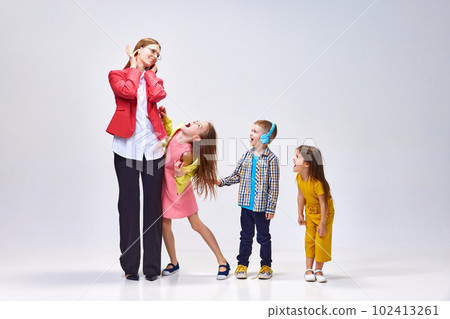 Rest needed. Portrait of young woman covering ears to avoid noisy kids shouting, playing around her against grey studio background Rest needed. Portrait of young woman covering ears to avoid noisy kids shouting, playing around her against grey studio background 102413261