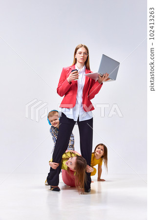 Portrait of businesswoman with laptop and little children playing around her with noise against grey studio background Portrait of businesswoman with laptop and little children playing around her with noise against grey studio background 102413263