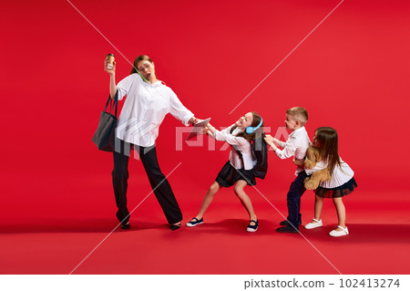 Portrait of businesswoman, mother talking on phone, little boy and girls, children pulling her hand, disturbing her against red studio background 102413274