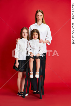 Portrait of beautiful family, mother and two little girls posing in same style clothes together against red studio background 102413298