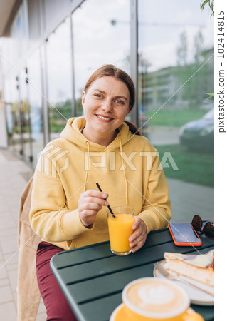 Happy woman sitting on the cafe terrace on the modern city street. 30s Person sitting at table and drinking drinks freshly squeezed orange fresh juice through straw in glass. Concept of easy breakfast 102414815