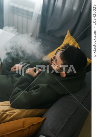 Bearded millennial or gen z man smoking hookah while relaxing on sofa at home - chill time and resting concept 102420029