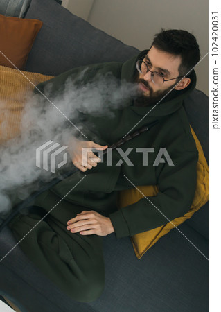 Bearded man is smoking hookah at home and blowing cloud of smoke - chill time and resting concept Bearded man is smoking hookah at home and blowing cloud of smoke - chill time and resting concept 102420031