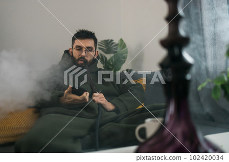 Bearded man is smoking hookah at home and blowing cloud of smoke - chill time and resting concept 102420034