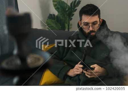 Bearded man is smoking hookah at home and blowing cloud of smoke - chill time and resting concept 102420035