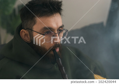 Bearded millennial or gen z man smoking hookah while relaxing on sofa at home - chill time and resting concept Bearded millennial or gen z man smoking hookah while relaxing on sofa at home - chill time and resting concept 102420044
