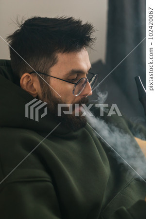 Bearded man is smoking hookah at home and blowing cloud of smoke - chill time and resting concept 102420067