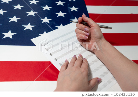 Female hands writing musical notes on a sheet of sheet music, against the background of the American flag, The concept of creating music Female hands writing musical notes on a sheet of sheet music, against the background of the American flag, The concept of creating music 102423130