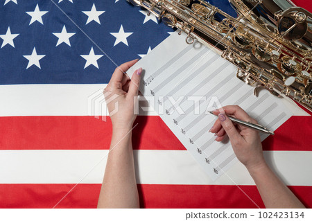 Female hands writing musical notes on a sheet of sheet music, against the background of the American flag, The concept of creating music Female hands writing musical notes on a sheet of sheet music, against the background of the American flag, The concept of creating music 102423134