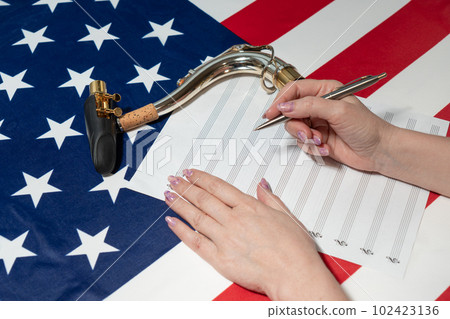 Female hands writing musical notes on a sheet of sheet music, against the background of the American flag, The concept of creating music Female hands writing musical notes on a sheet of sheet music, against the background of the American flag, The concept of creating music 102423136