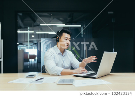 Portrait of a young Asian man in a headset. Sits in the office at the laptop and consults, talks through a video call from the laptop. Portrait of a young Asian man in a headset. Sits in the office at the laptop and consults, talks through a video call from the laptop. 102423154