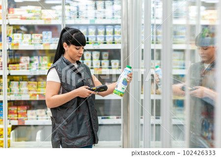 A young Caucasian woman working as a Manager in a supermarket checks the expiration date of milk in a bottle, using an electronic device. Business and technology concepts 102426333