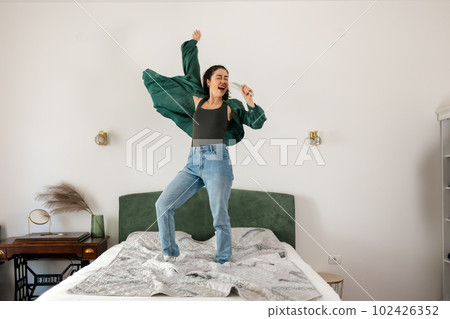 Winning casino bet. Young Caucasian happy woman is singing in hairbrush and dancing on bed. Concept of dance and vocals learning Winning casino bet. Young Caucasian happy woman is singing in hairbrush and dancing on bed. Concept of dance and vocals learning 102426352