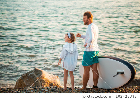 Man and preschool girl standing on the beach with sup board. Ocean's surface on the background. Summer active sport at the sea. Back view 102426383