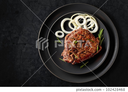 Grilled ribeye beef steak with rosemary and marinated onion on a black stone table. Grilled ribeye beef steak with rosemary and marinated onion on a black stone table. 102428468