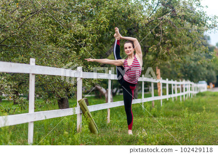 Full length portrait of flexible strong young girl stretching her leg, practicing yoga, having workout outdoor, raising leg in air, doing split stretch one foot, looking smiling away. Full length portrait of flexible strong young girl stretching her leg, practicing yoga, having workout outdoor, raising leg in air, doing split stretch one foot, looking smiling away. 102429118