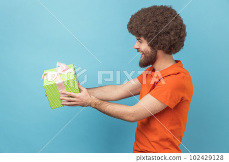 Side view of man with Afro hairstyle wearing orange T-shirt giving gift, congratulating on birthday and offering surprise, holiday bonus wrapped in box. Indoor studio shot isolated on blue background. Side view of man with Afro hairstyle wearing orange T-shirt giving gift, congratulating on birthday and offering surprise, holiday bonus wrapped in box. Indoor studio shot isolated on blue background. 102429128