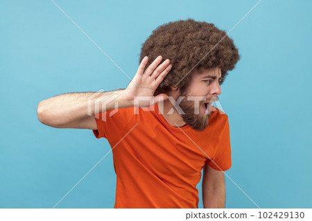 Portrait of man with Afro hairstyle in T-shirt holding hand near ear trying to listen quiet conversation, overhearing gossip, having hearing problems. Indoor studio shot isolated on blue background. Portrait of man with Afro hairstyle in T-shirt holding hand near ear trying to listen quiet conversation, overhearing gossip, having hearing problems. Indoor studio shot isolated on blue background. 102429130