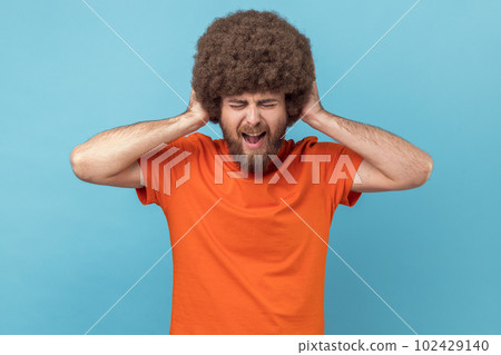 Portrait of man with Afro hairstyle covering ears, cannot focus in crowded place, feels discomfort over noise, being troubled to listen advice. Indoor studio shot isolated on blue background. 102429140