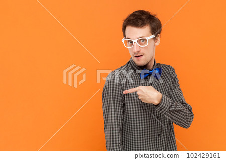 Portrait of man nerd pointing away on copy space, discusses amazing promo, gives way or direction, wearing shirt with blue bow tie and white glasses. Indoor studio shot isolated on orange background. Portrait of man nerd pointing away on copy space, discusses amazing promo, gives way or direction, wearing shirt with blue bow tie and white glasses. Indoor studio shot isolated on orange background. 102429161