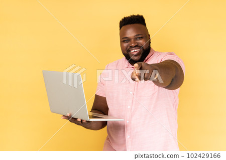 Portrait of optimistic bearded man wearing pink shirt holding laptop, pointing finger to camera and smiling, choosing you, freelance job advertisement. Indoor studio shot isolated on yellow background 102429166