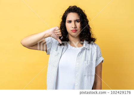 Portrait of woman with dark wavy hair criticizing bad quality with thumbs down displeased grimace, showing dislike gesture, expressing disapproval. Indoor studio shot isolated on yellow background. Portrait of woman with dark wavy hair criticizing bad quality with thumbs down displeased grimace, showing dislike gesture, expressing disapproval. Indoor studio shot isolated on yellow background. 102429176