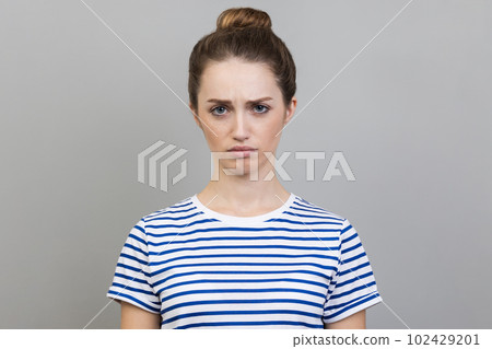 Portrait of sad upset young adult woman wearing striped T-shirt expressing sadness, hearing bad news, being in bad mood. Indoor studio shot isolated on gray background. Portrait of sad upset young adult woman wearing striped T-shirt expressing sadness, hearing bad news, being in bad mood. Indoor studio shot isolated on gray background. 102429201