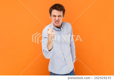 Portrait of crazy man shows fuck you sign looking with poker face being vulgar and has quarrel with someone, wearing light blue shirt. Indoor studio shot isolated on orange background. 102429235