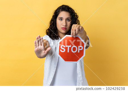 Portrait of strict woman with dark wavy hair holding red stop sign looking at camera with ban palm gesture, has strict expression, prohibition. Indoor studio shot isolated on yellow background. 102429238