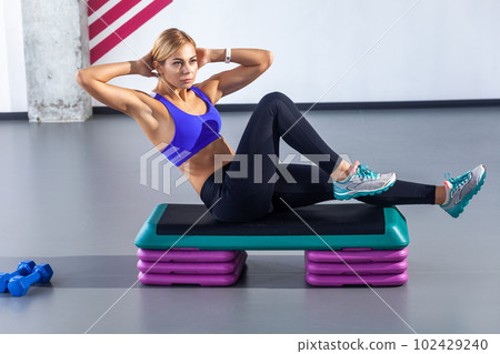Side view portrait of sporty beautiful woman with blonde hair wearing tight sportswear sitting on step platform and doing press aerobic, having work out in fitness club. Indoor shot. 102429240