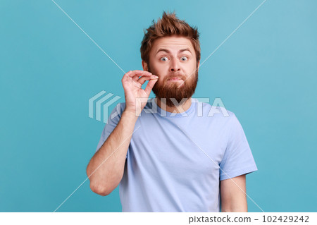 I don't tell anyone. Portrait of handsome young adult bearded man zipping mouth, keeping secret information, looking at camera. Indoor studio shot isolated on blue background. 102429242