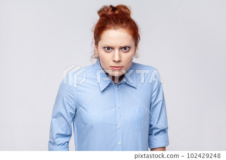 Portrait of angry aggressive ginger woman wearing blue shirt looking at camera with anger and hate, being in bad nervous mood. Indoor studio shot isolated on gray background. Portrait of angry aggressive ginger woman wearing blue shirt looking at camera with anger and hate, being in bad nervous mood. Indoor studio shot isolated on gray background. 102429248