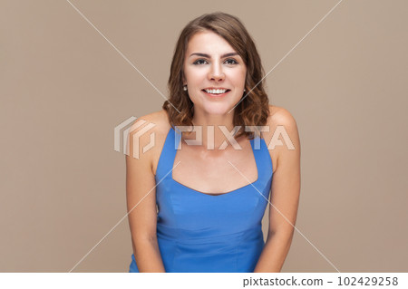 Portrait of smiling positive happy woman with wavy hair looking at camera with toothy smile, being in good mood, being glad, wearing blue dress. Indoor studio shot isolated on light brown background. Portrait of smiling positive happy woman with wavy hair looking at camera with toothy smile, being in good mood, being glad, wearing blue dress. Indoor studio shot isolated on light brown background. 102429258