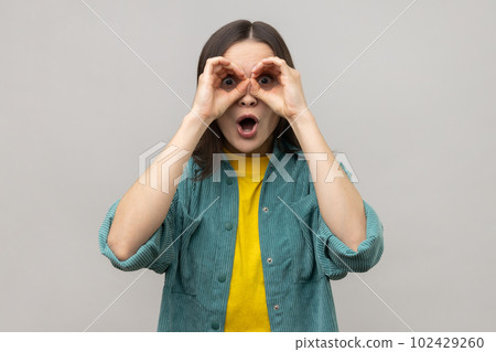 Shocked attractive young woman standing with hands on eyes binoculars gesture and looking at camera with surprised face, wearing casual style jacket. Indoor studio shot isolated on gray background. Shocked attractive young woman standing with hands on eyes binoculars gesture and looking at camera with surprised face, wearing casual style jacket. Indoor studio shot isolated on gray background. 102429260