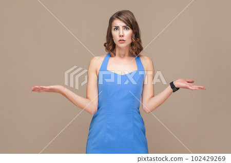 Portrait of puzzled confused young adult woman with wavy hair standing shrugging shoulders, don't know what to do, wearing blue dress. Indoor studio shot isolated on light brown background. Portrait of puzzled confused young adult woman with wavy hair standing shrugging shoulders, don't know what to do, wearing blue dress. Indoor studio shot isolated on light brown background. 102429269
