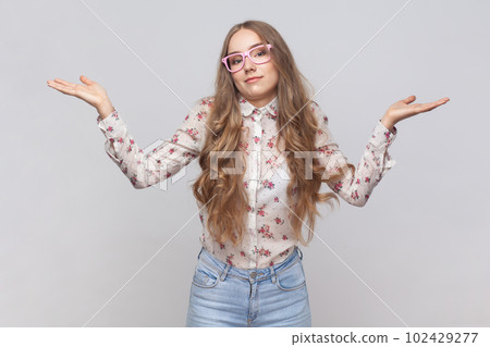 I don't know. Portrait of confused beautiful woman in glasses with wavy blond hair standing, raised arms, confused and thinking. Indoor studio shot isolated on gray background. 102429277